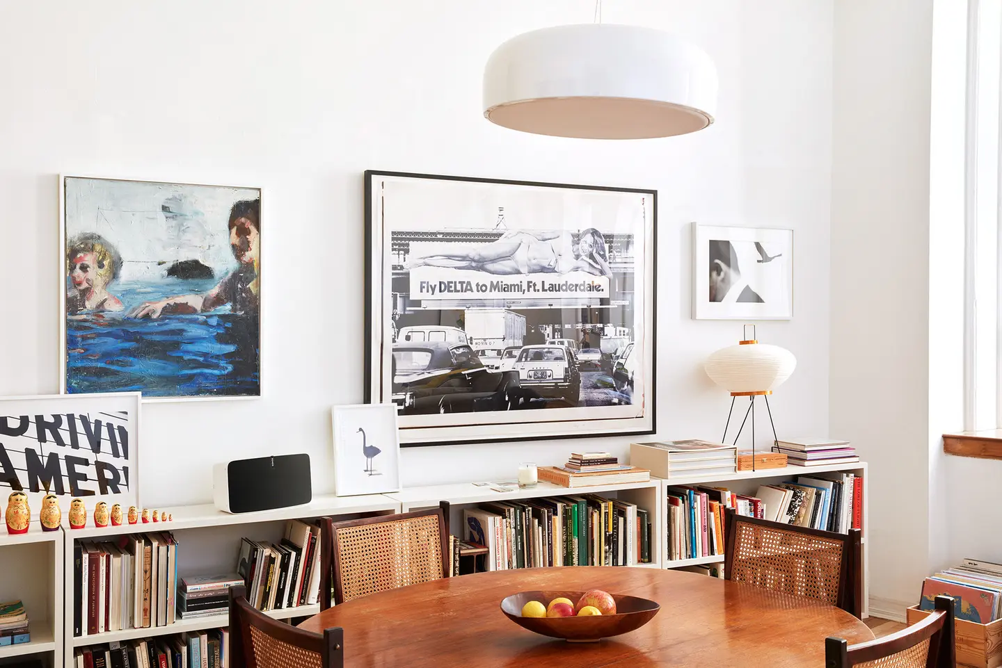 Ein runder Esstisch mit Obstschale, umgeben von Stühlen, vor Regalen mit Büchern und mehreren Kunstwerken an der Wand.