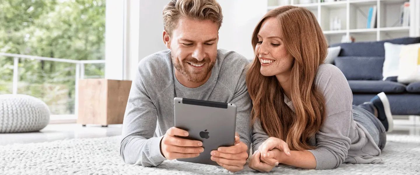 Ein Mann und eine Frau liegen nebeneinander auf einem Teppich und schauen gemeinsam auf ein Tablet.