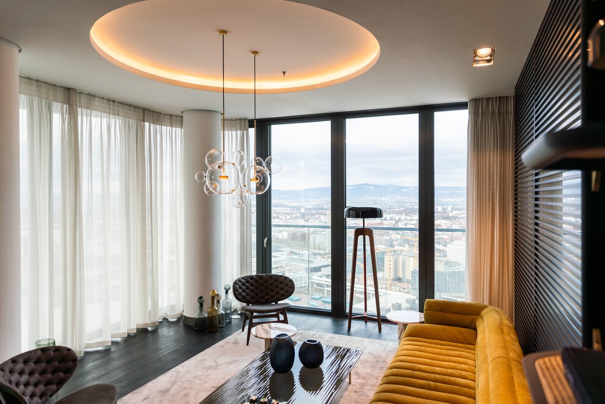 Moderne Wohnzimmer mit gelbem Sofa, beigem Teppich, Beleuchtungskörper und großen Fenstern.