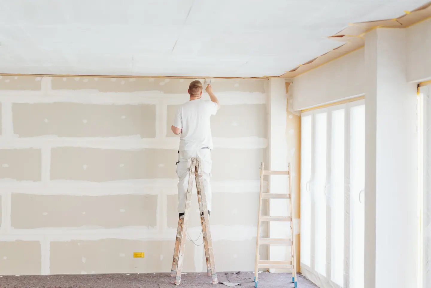 Mann auf einer Leiter, der die Decke eines Raumes streicht, während eine unvollendete Wand sichtbar ist.