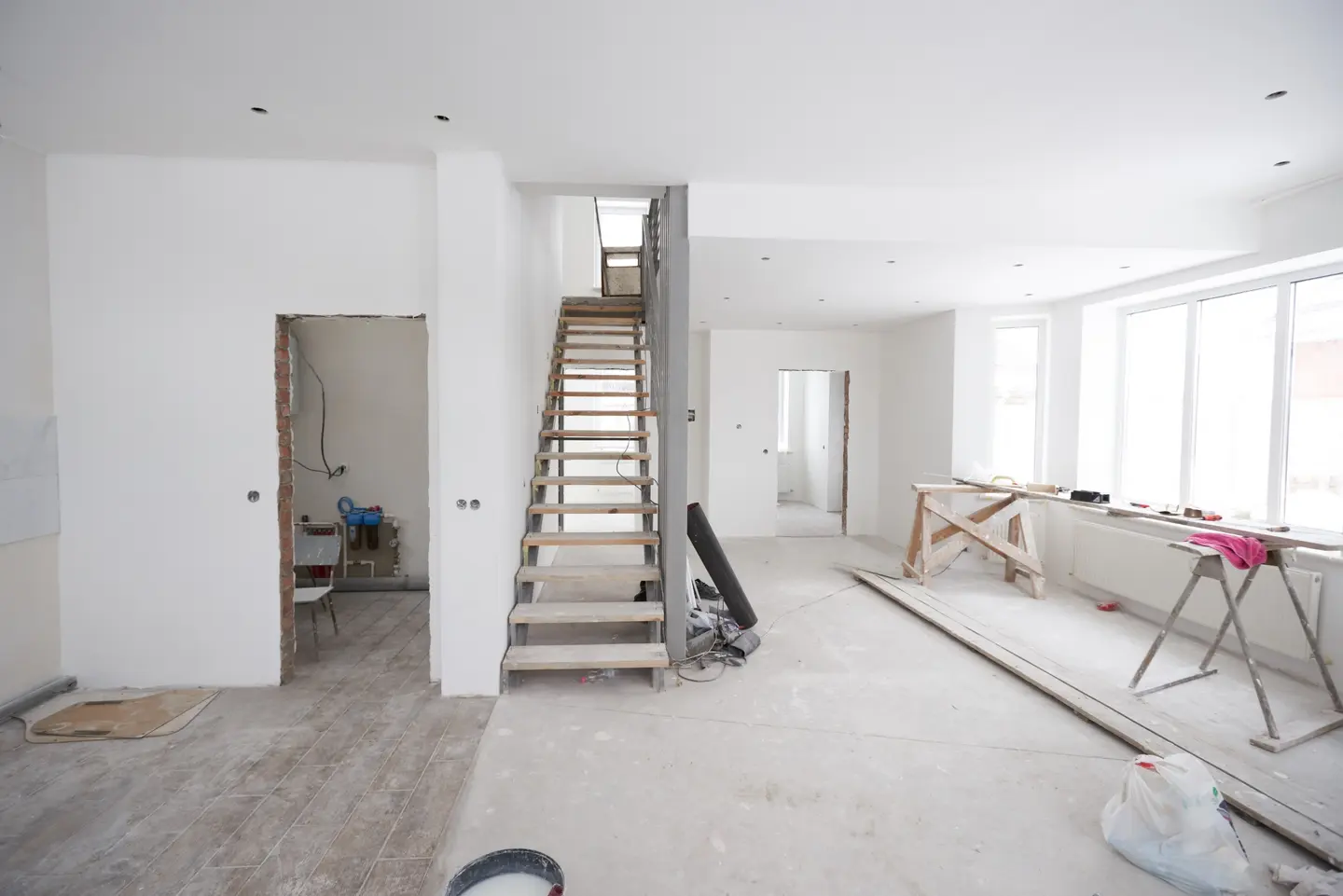 Ein unfertiger Raum mit einer Treppe, zwei Türen und Baugeräten, umgeben von unvollendeten Wänden und Bodenbelag.