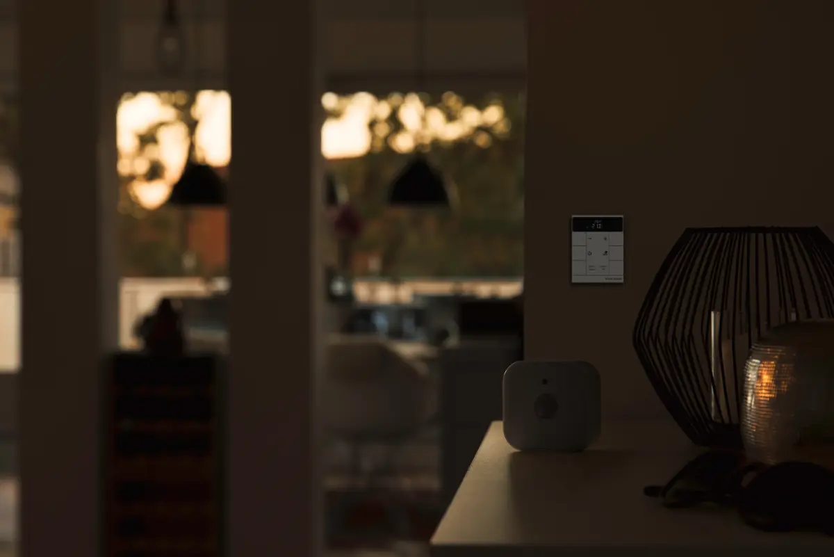 Een smart home-bedieningspaneel aan de muur, naast een decoratieve lamp en een klein apparaat op een tafel.  