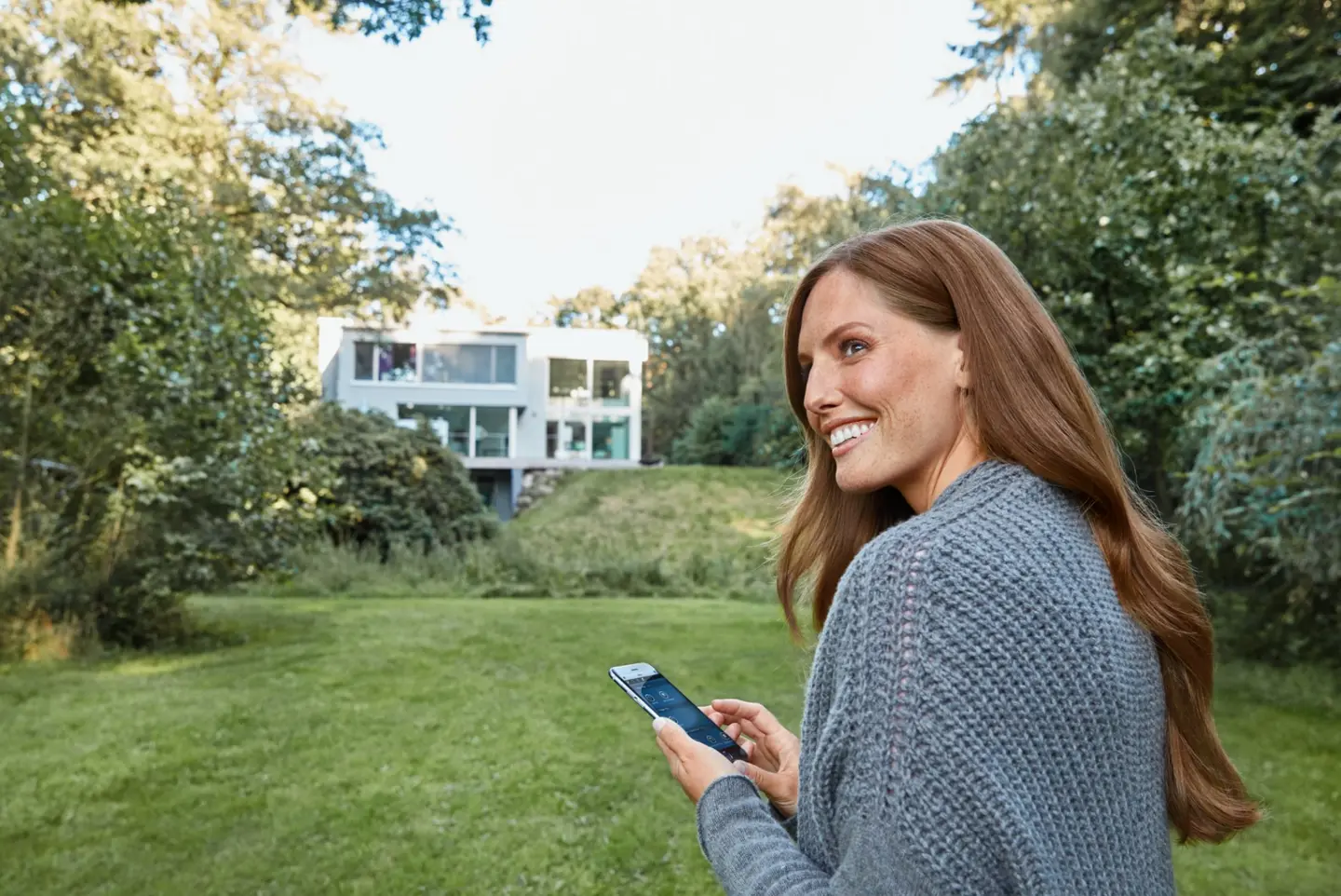 Eine Frau mit langen Haaren und einem grauen Pullover, die auf ihr Smartphone schaut, steht im Garten.