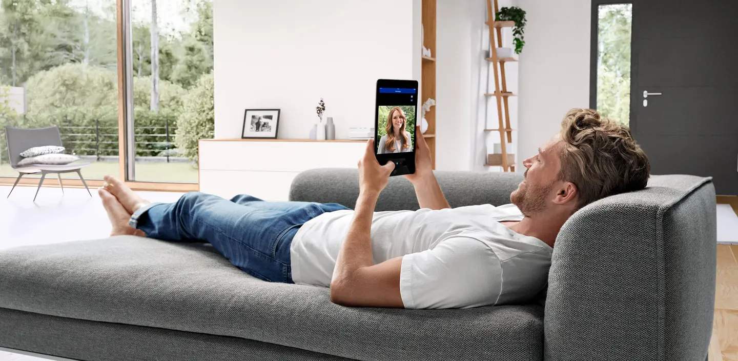 Man ligt op de bank en houdt de deurcommunicatie app vast op zijn tablet