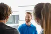Drei Personen stehen sich gegenüber; ein Mann in einem blauen Poloshirt lächelt freundlich.