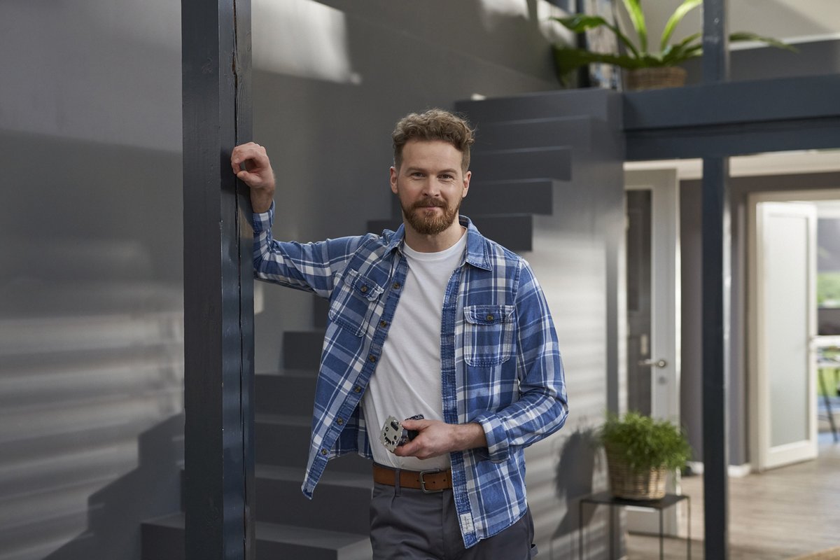 Ein Mann steht an einer Wand und hält einen Gegenstand in der Hand, gekleidet in ein kariertes Hemd und T-Shirt.