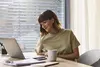 Eine Frau sitzt an einem Tisch mit einem Laptop, Blick auf den Bildschirm, Tasse und Notizblock daneben.