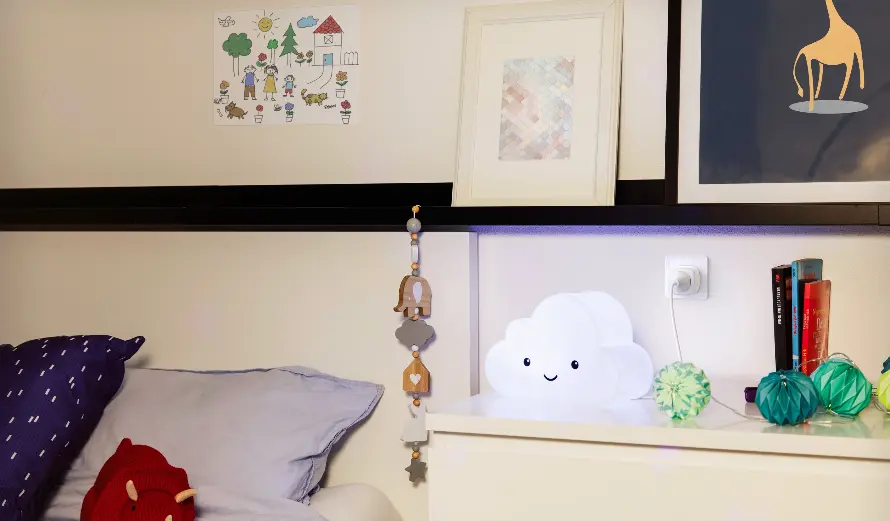 Nachtkastje met een wolkenlamp, een boek en decoratieve elementen; op het bed ligt een kussen.