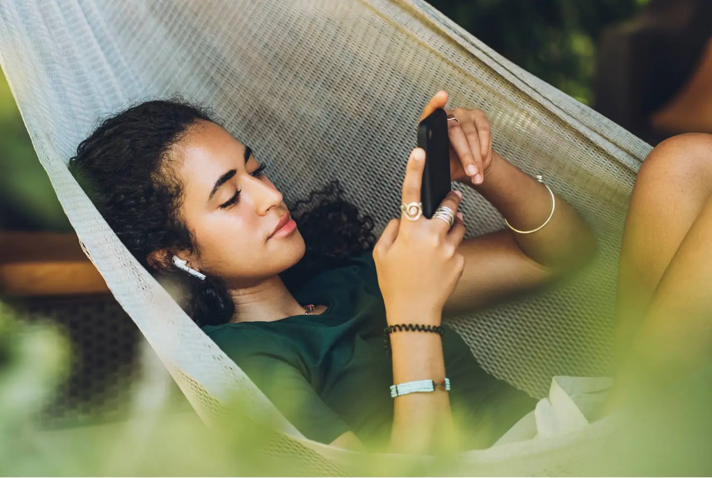 Eine Person liegt entspannt in einer Hängematte und schaut auf ihr Smartphone, während sie kabellose Ohrhörer trägt.