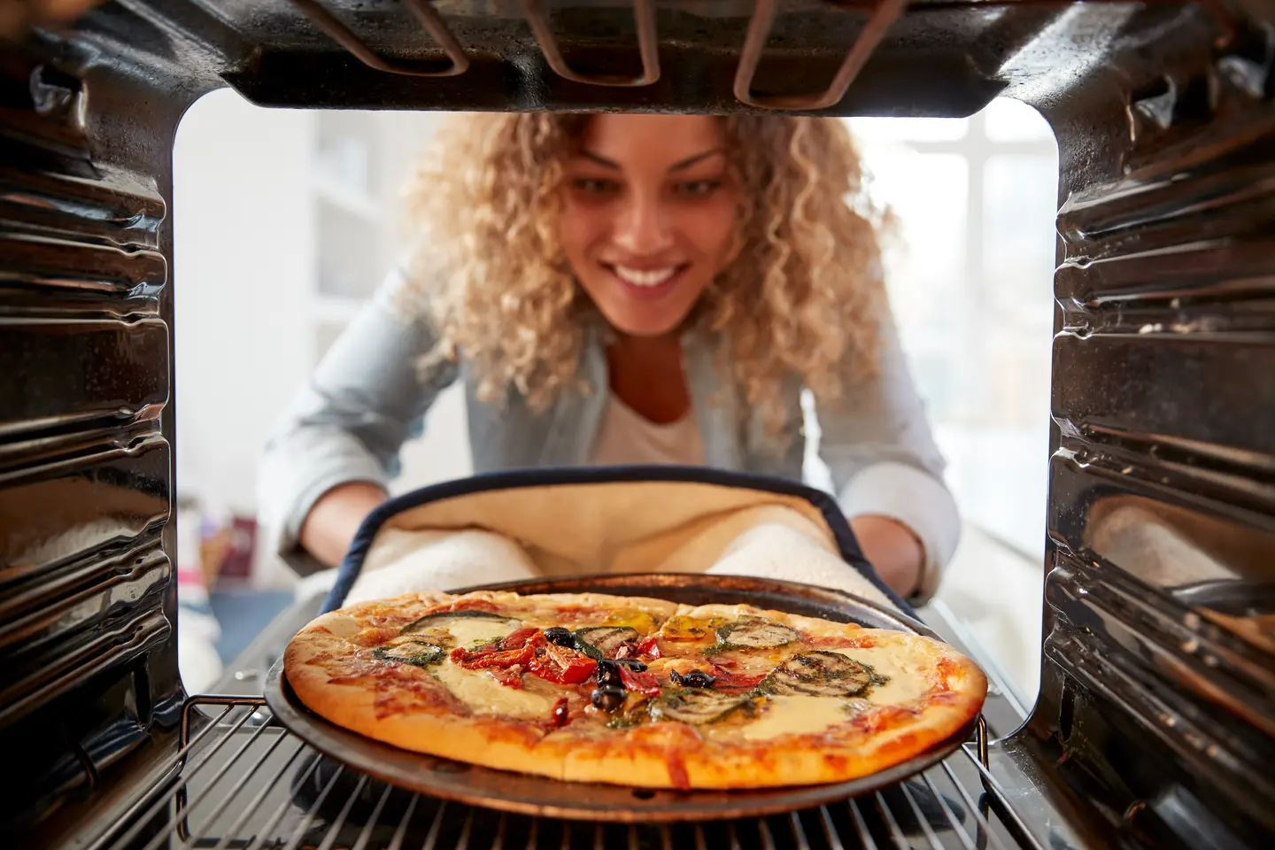 Eine Person mit lockigem Haar blickt freudig auf eine frisch gebackene Pizza im Ofen.