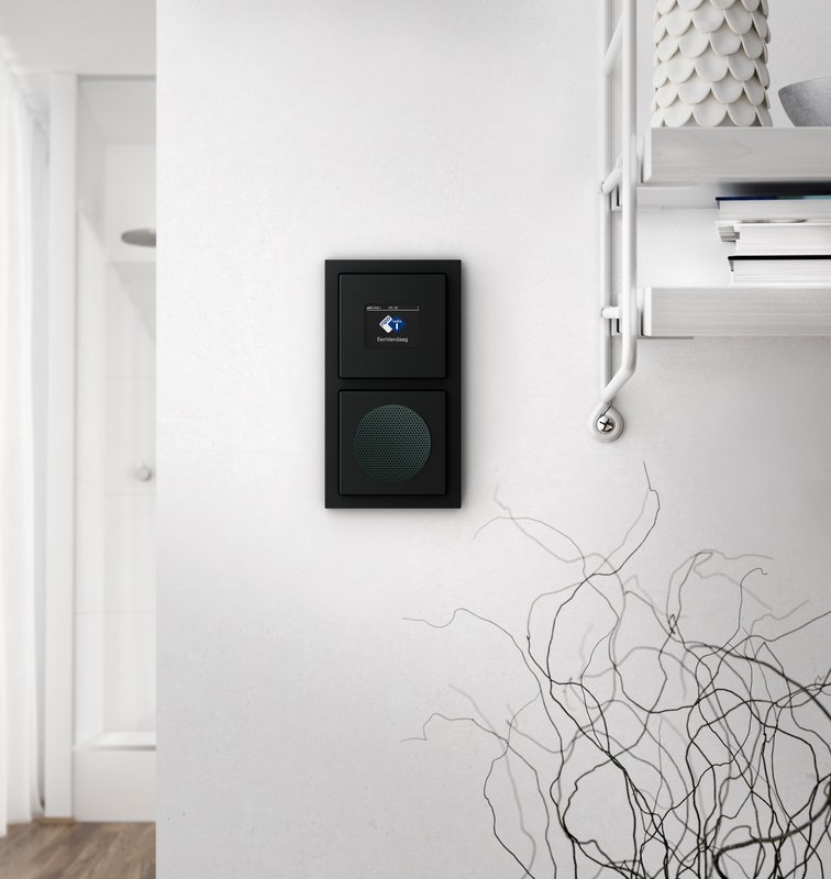 Schwarzes Wandgerät mit Lautsprecher und blauen LED-Anzeigen, montiert an einer weißen Wand.