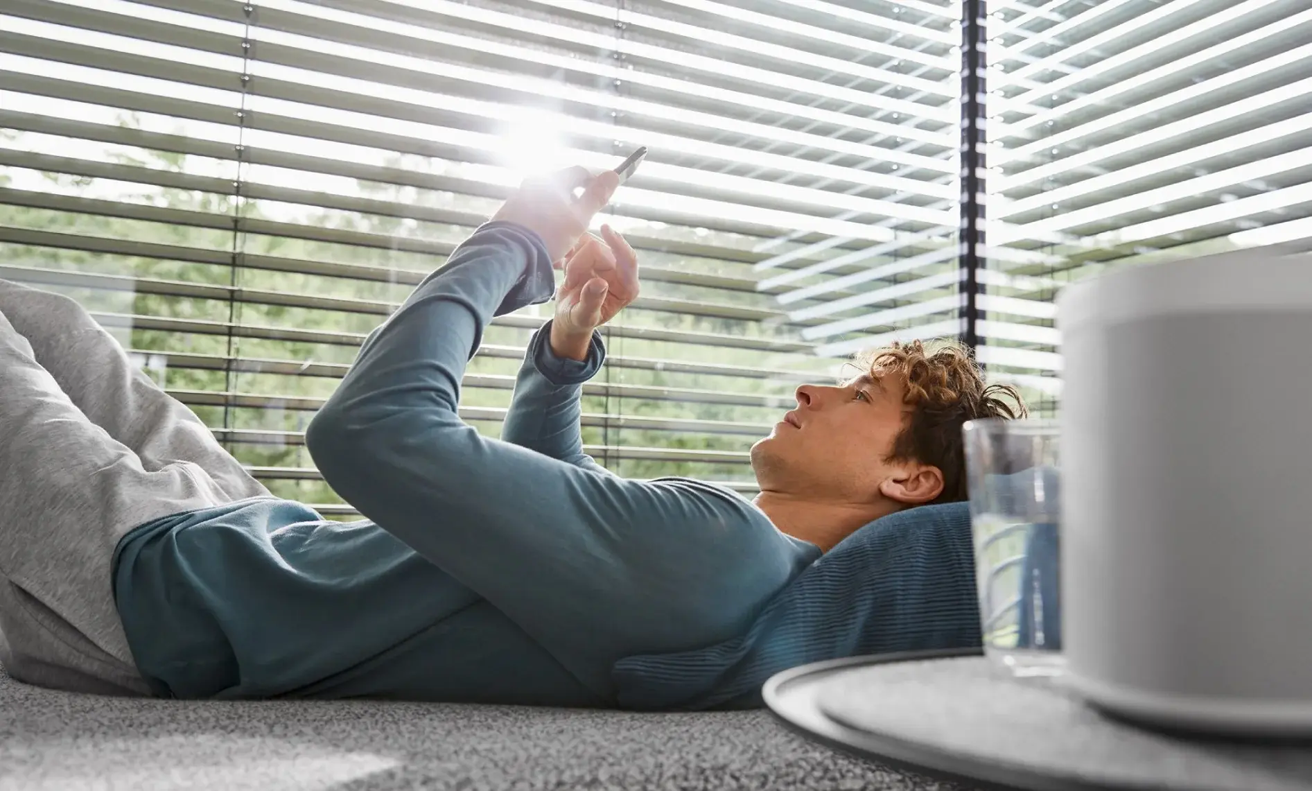 Junger Mann liegt auf einem Sofa und schaut auf sein Smartphone, während Sonnenlicht durch die Jalousien scheint.
