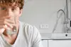 Ein Mann mit lockigem Haar trinkt Wasser aus einem Glas, während eine moderne Küchenspüle und Kräuterpflanze im Hintergrund zu sehen sind.