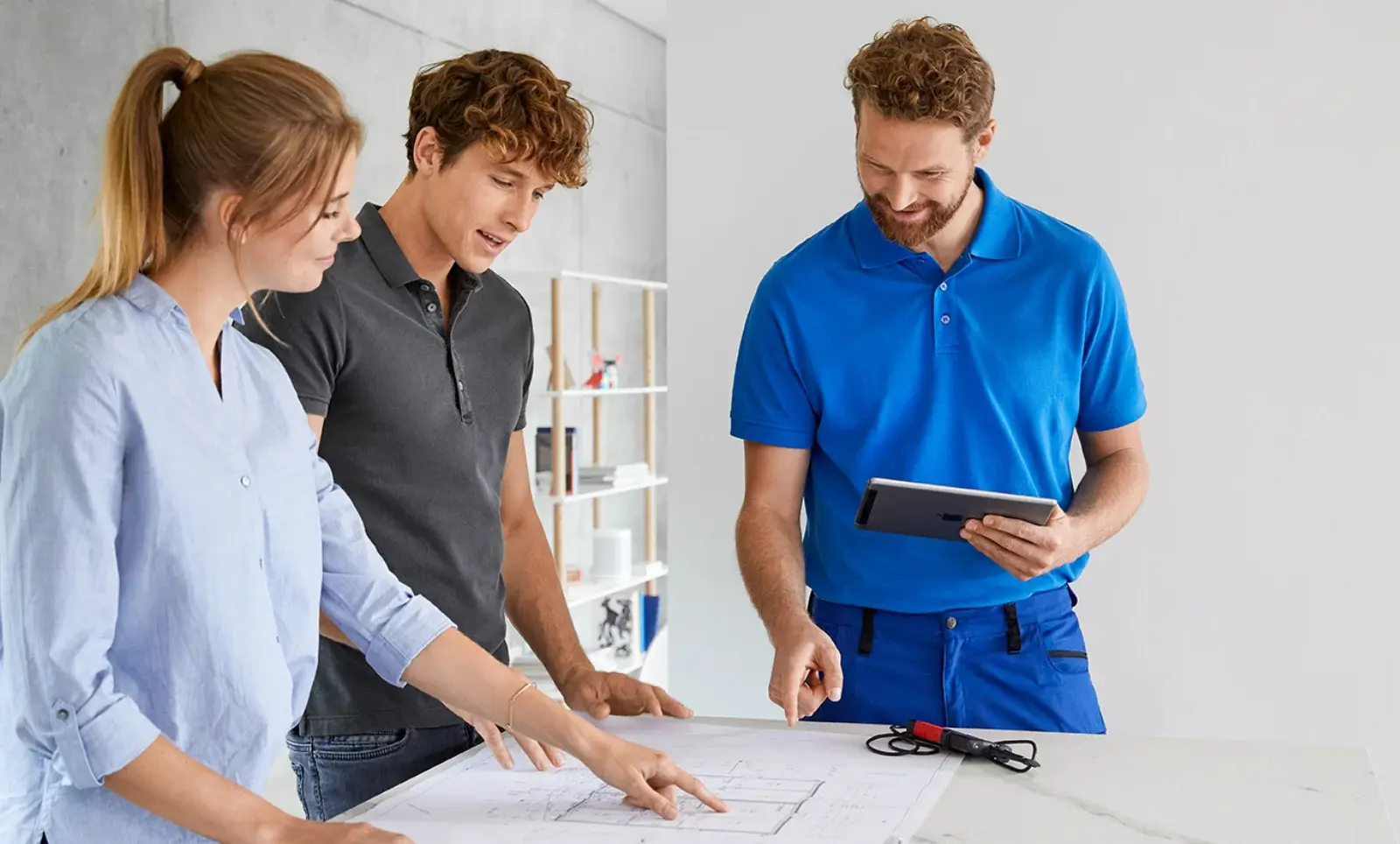 Drei Personen besprechen Baupläne an einem Tisch, während einer von ihnen ein Tablet in der Hand hält.