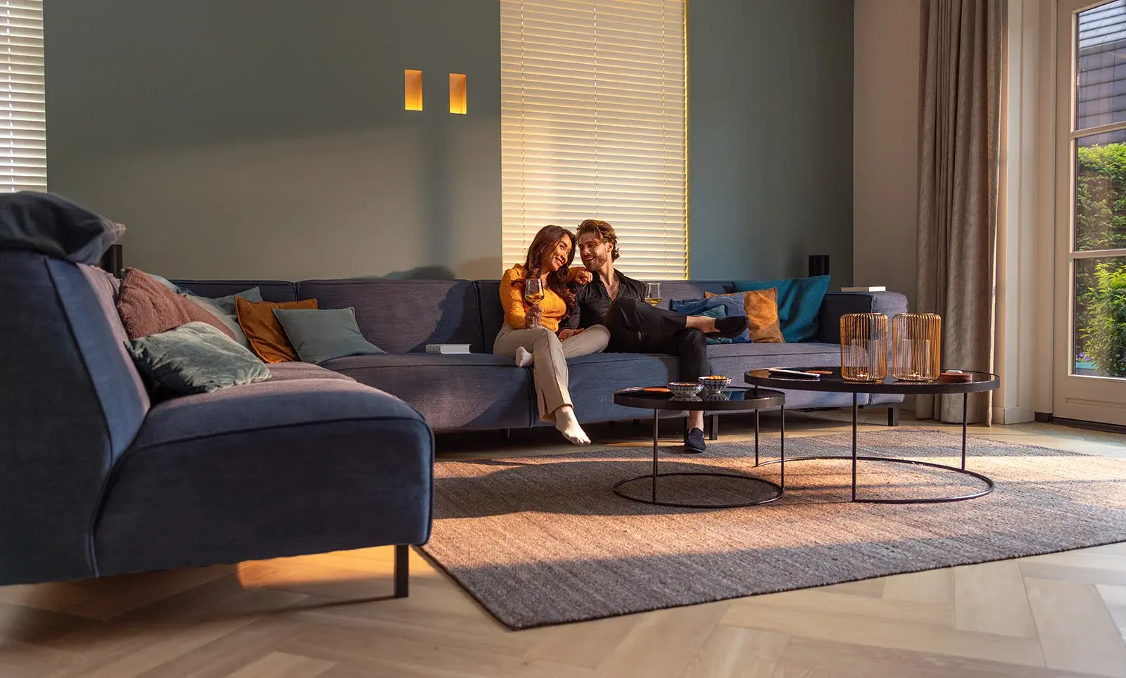 Ein Paar sitzt gemütlich auf einer Couch, umgeben von Kissen und einem Tisch mit Dekoartikeln in einem modernen Wohnzimmer.