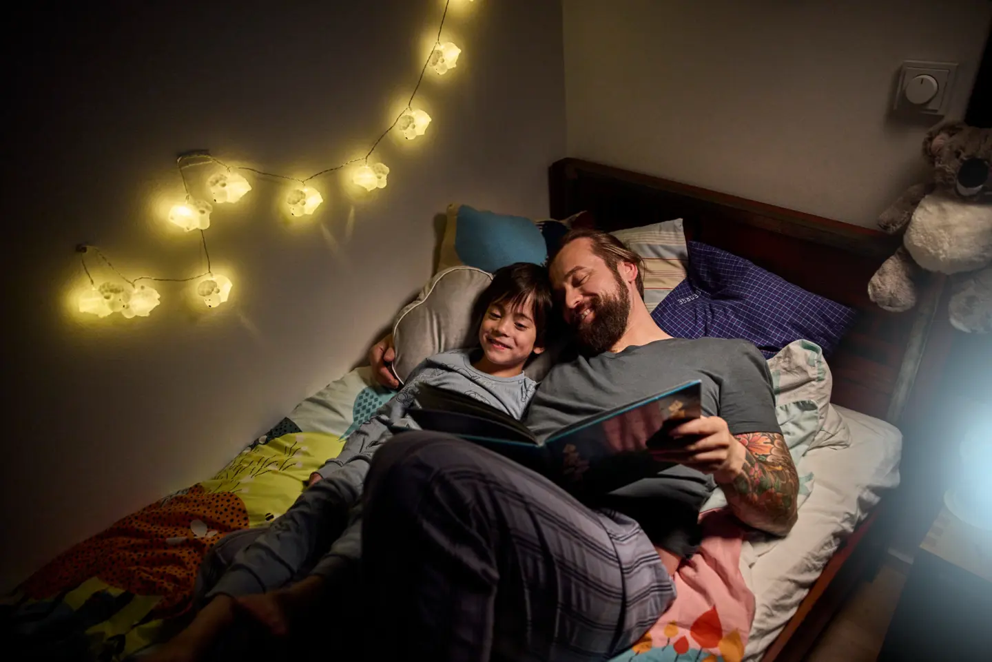 Een vader en kind lezen samen in bed, omringd door zacht licht en een gezellige sfeer. 