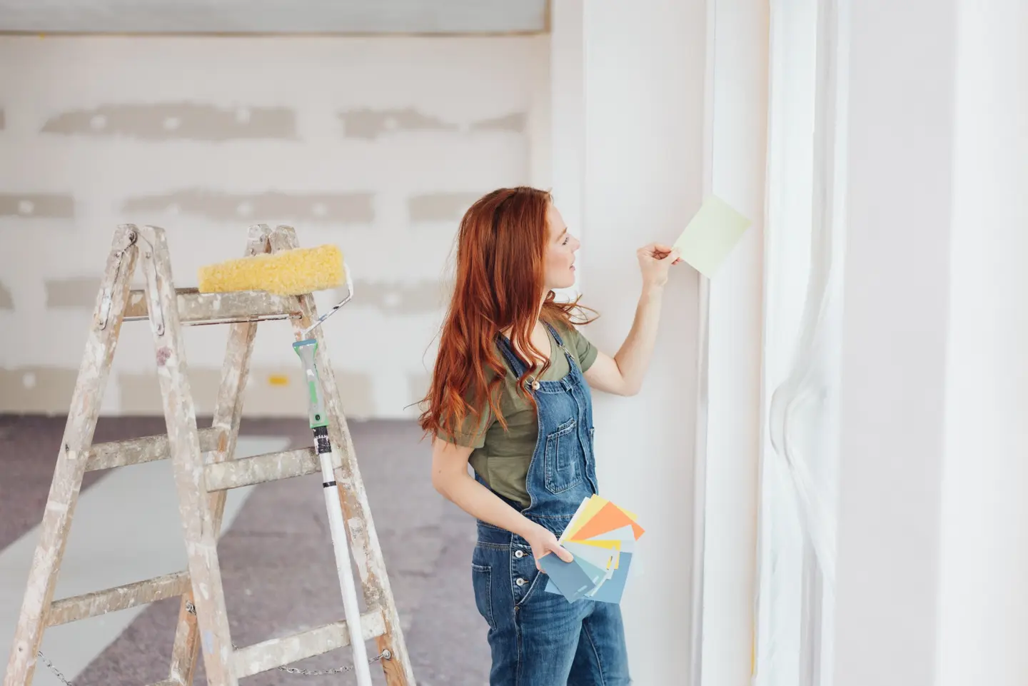 Frau mit roten Haaren, die Farbmuster an ein Fenster hält, neben einer Leiter mit Farbroller.